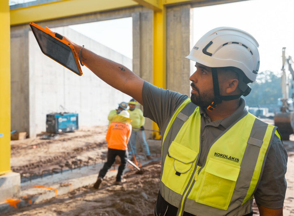 Man in safety vest holds tablet at construction site.