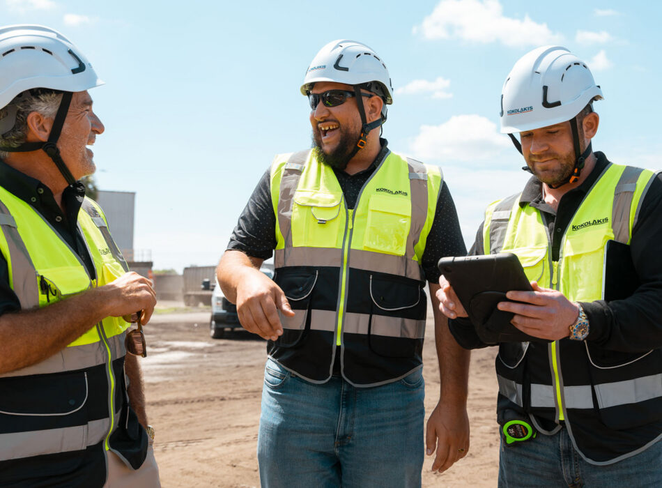 Three workers in safety gear look at a tablet outside.
