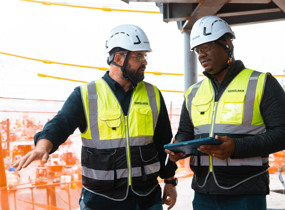 Two construction workers in safety gear at worksite with tablet.