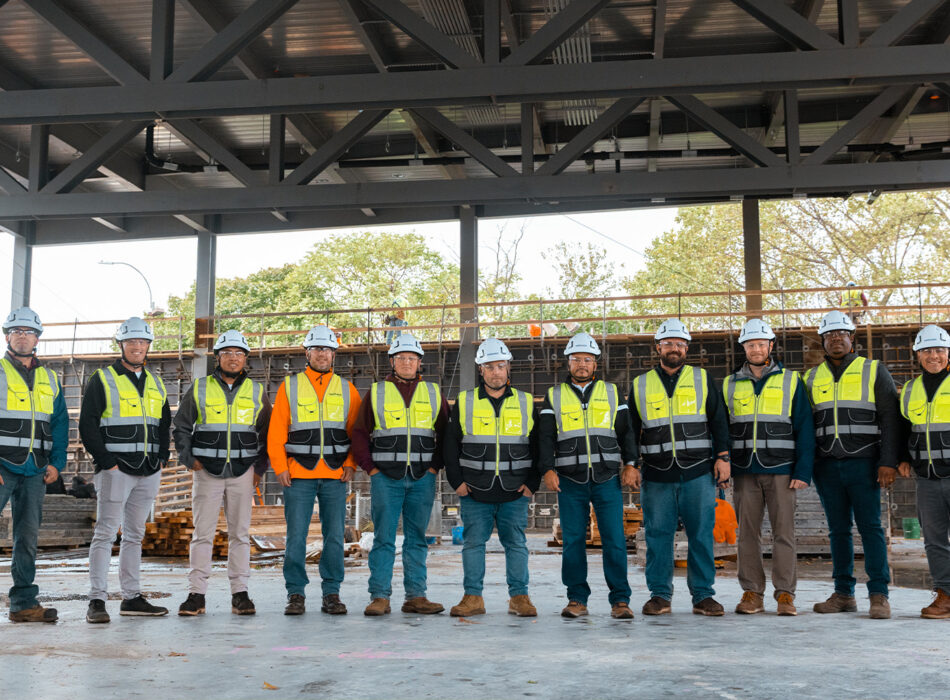 Eleven workers in safety gear line up at construction site.
