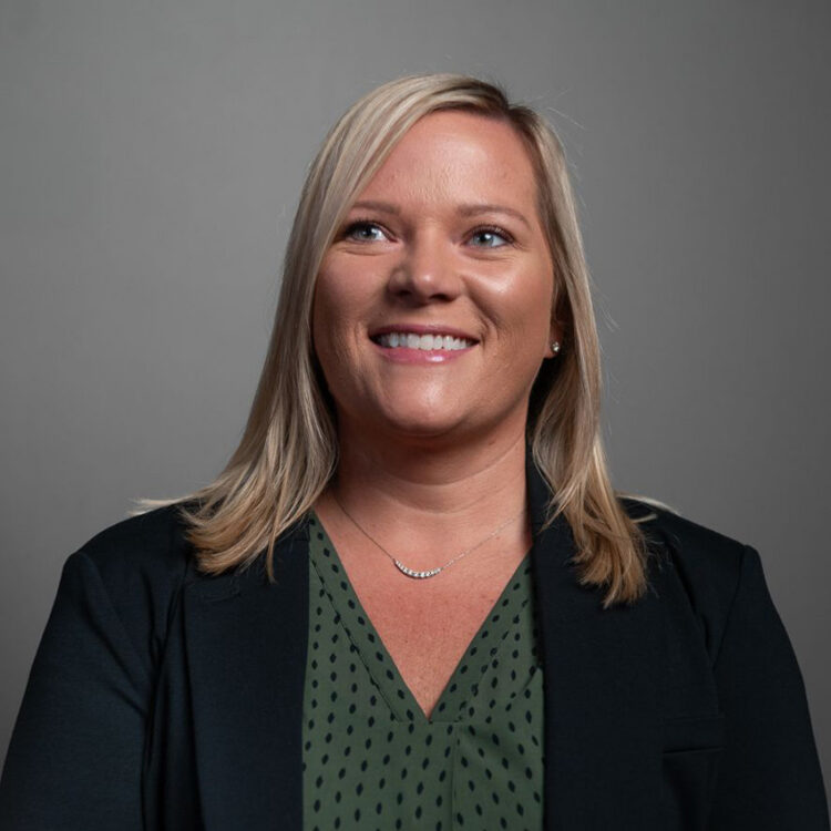 Blonde woman in dark blazer smiles before gray background.