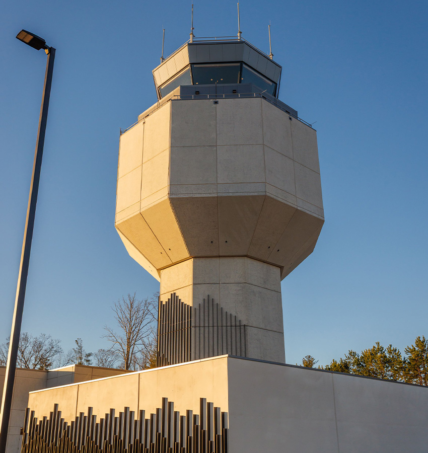 Concrete airport tower viewed from below, blue sky, trees.