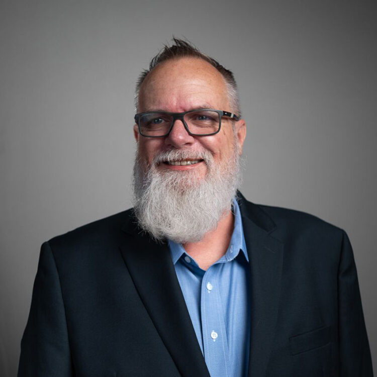Smiling man with glasses and white beard in blue shirt.