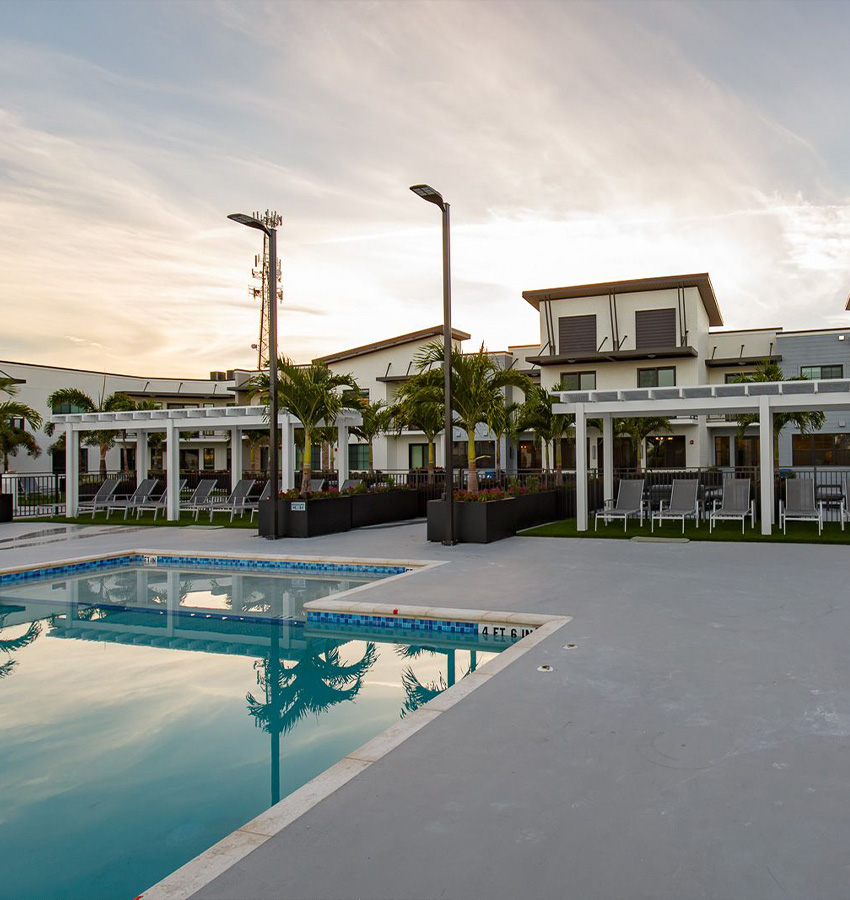 Pool, lounge chairs, and palm trees before modern buildings at sunset.