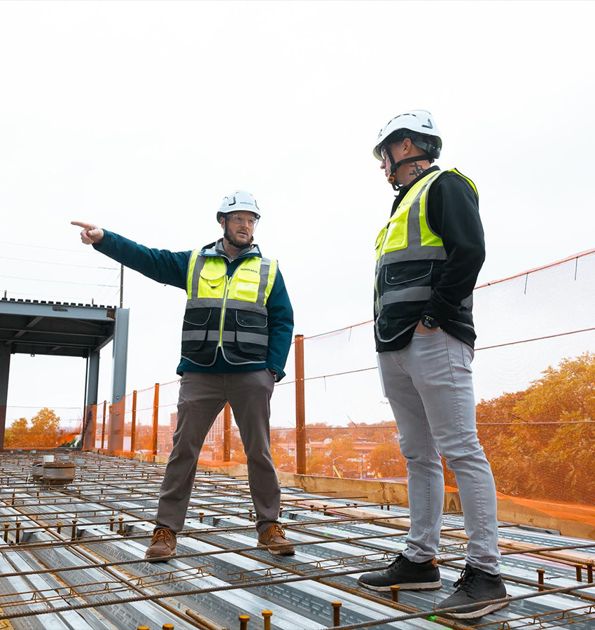 Two construction workers talk amid steel bars on site.