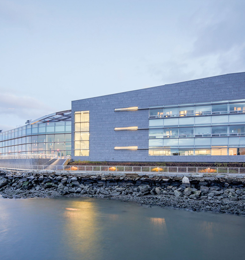 Modern illuminated building by rocky shore and calm water.