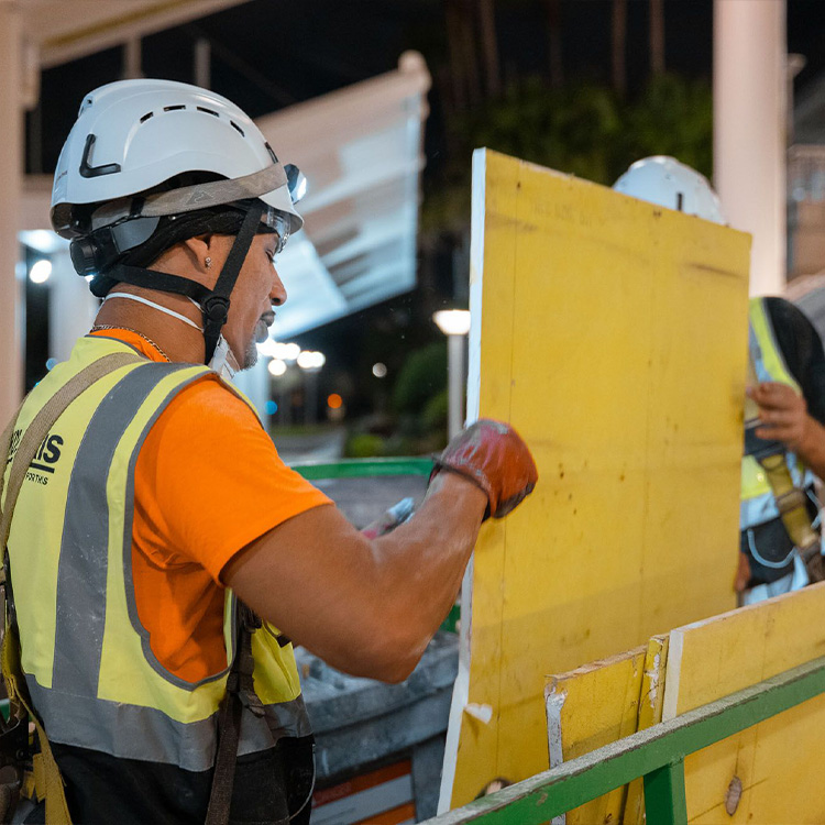 Worker in helmet moves yellow panel; another worker behind.