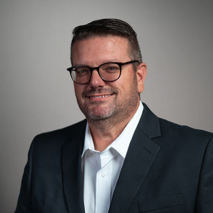 Smiling man in glasses, dark suit, white shirt, gray background.