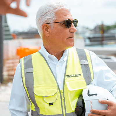 Older man in safety vest holds hard hat at site.