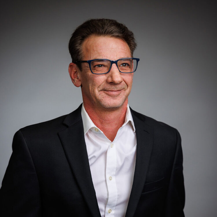 Smiling man in suit and glasses against gray background.