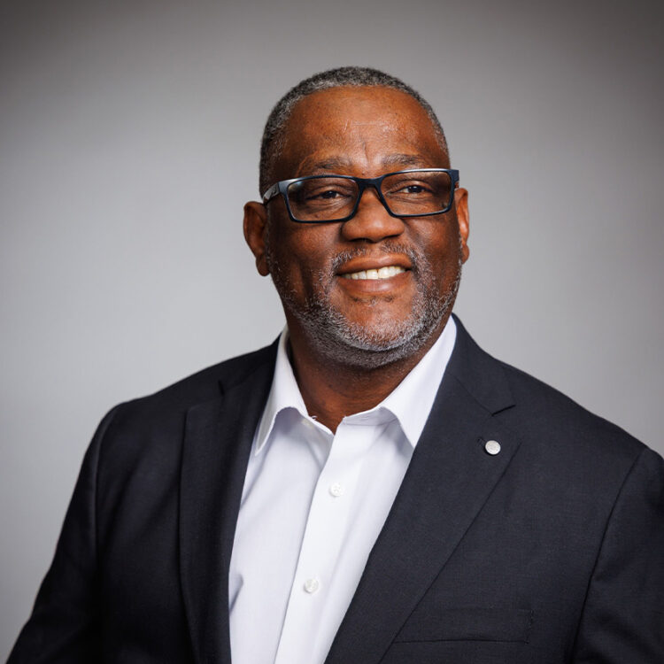 Smiling man in glasses and dark suit against gray background.