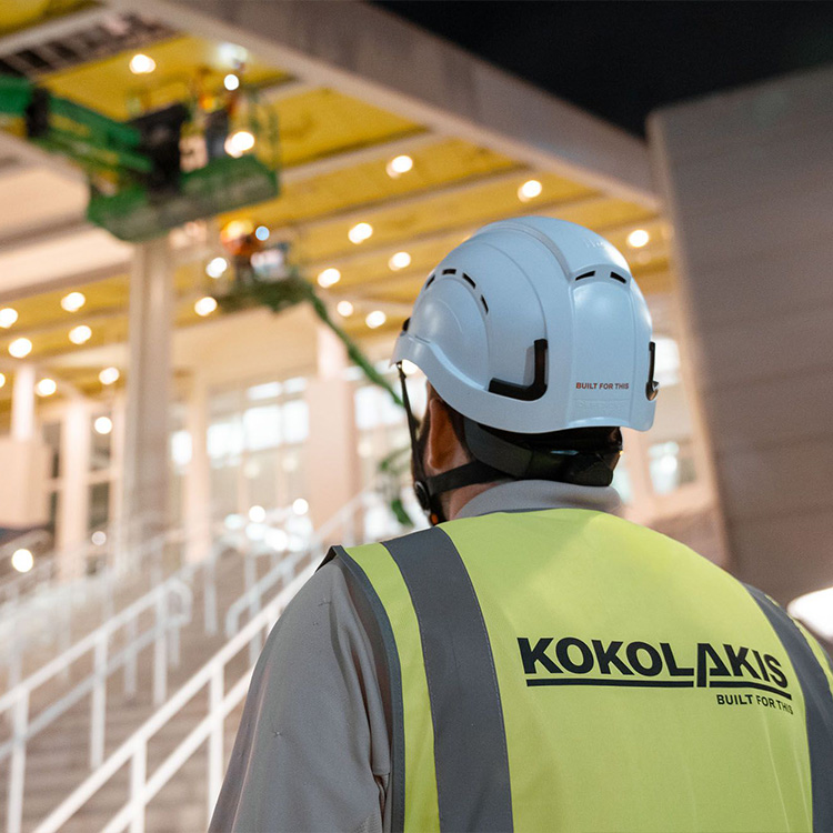 Worker in Kokolakis vest watches lift by building at night.