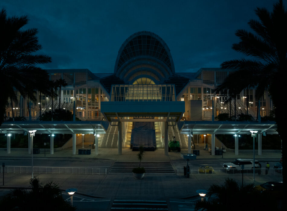 Modern glass building at night, palm trees, cloudy sky.