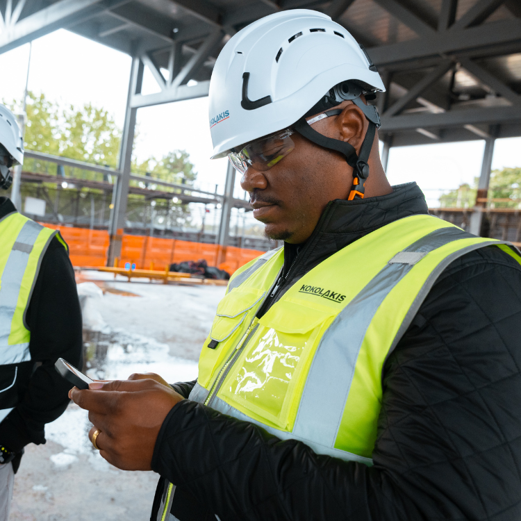Worker in safety gear uses smartphone at construction site.
