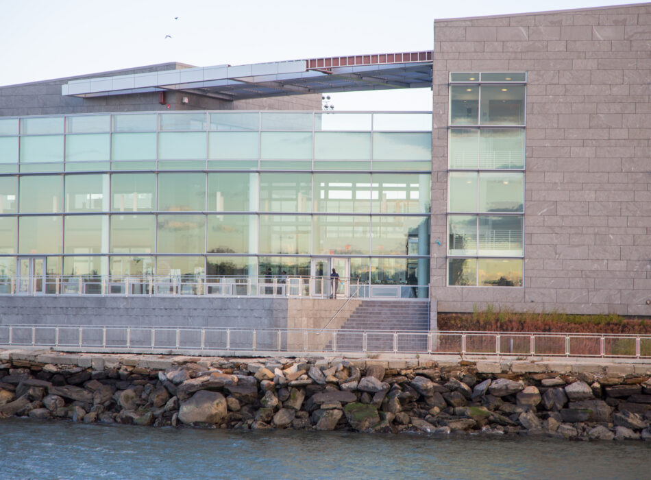 Modern glass building by rocky shore with water in front.