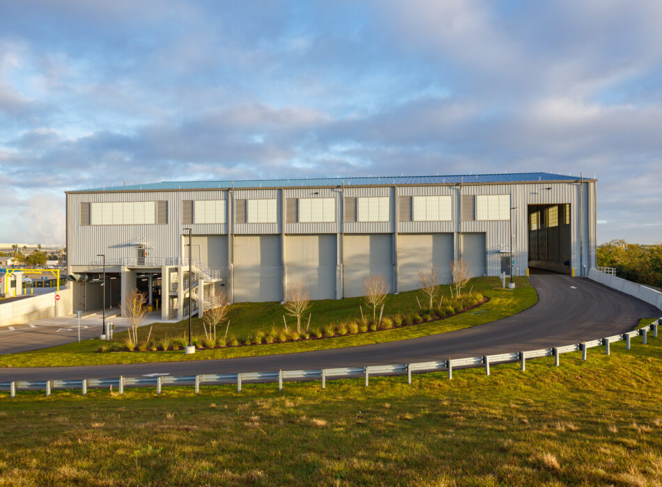 Gray warehouse with garage doors, ramp, grass, cloudy sky.