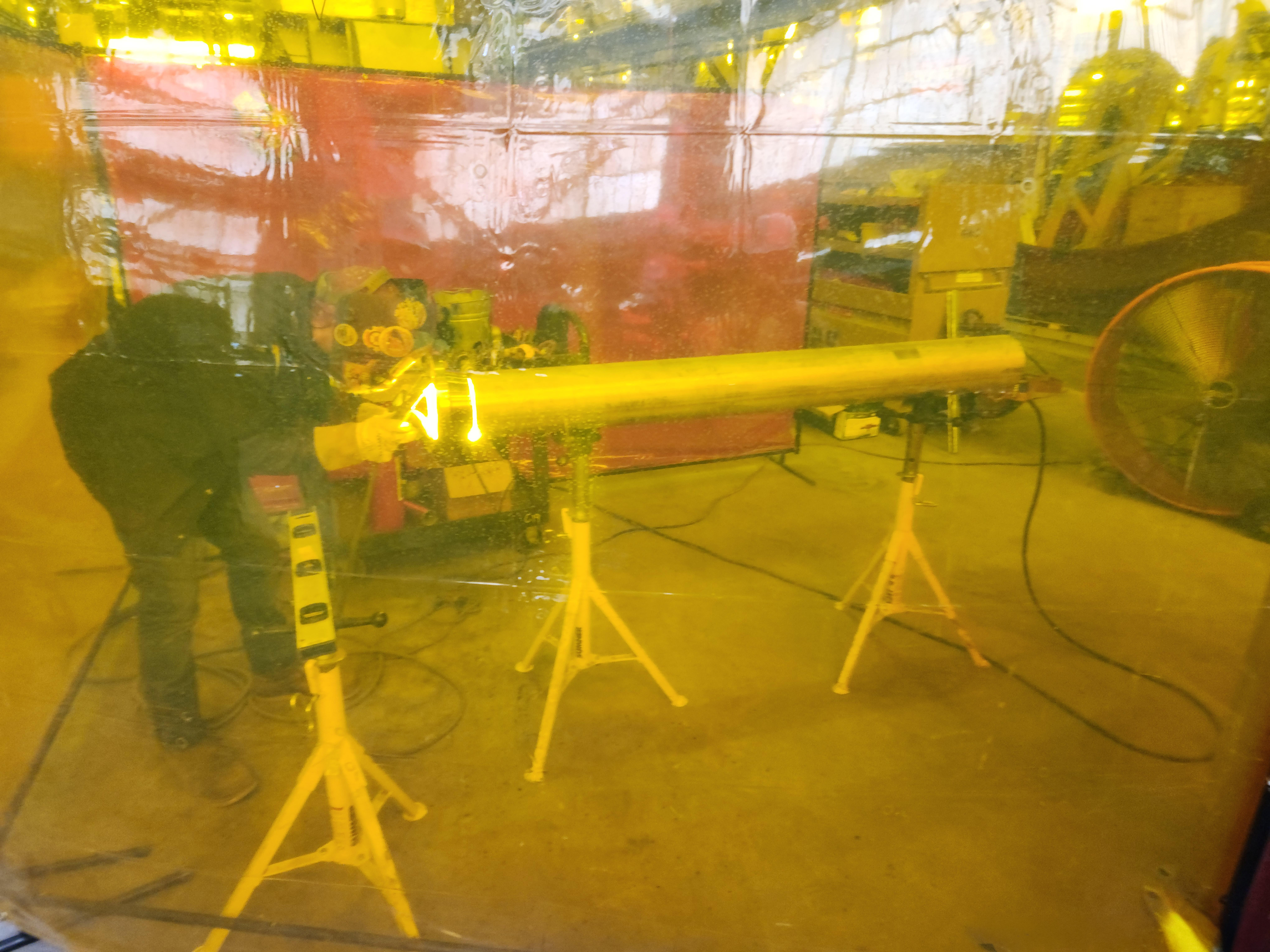 Person welding pipe in workshop behind yellow safety screen.