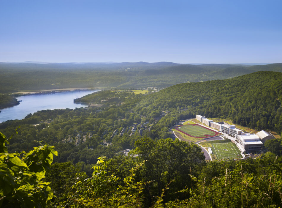 Building complex, sports fields, forested hills, lake, clear sky.