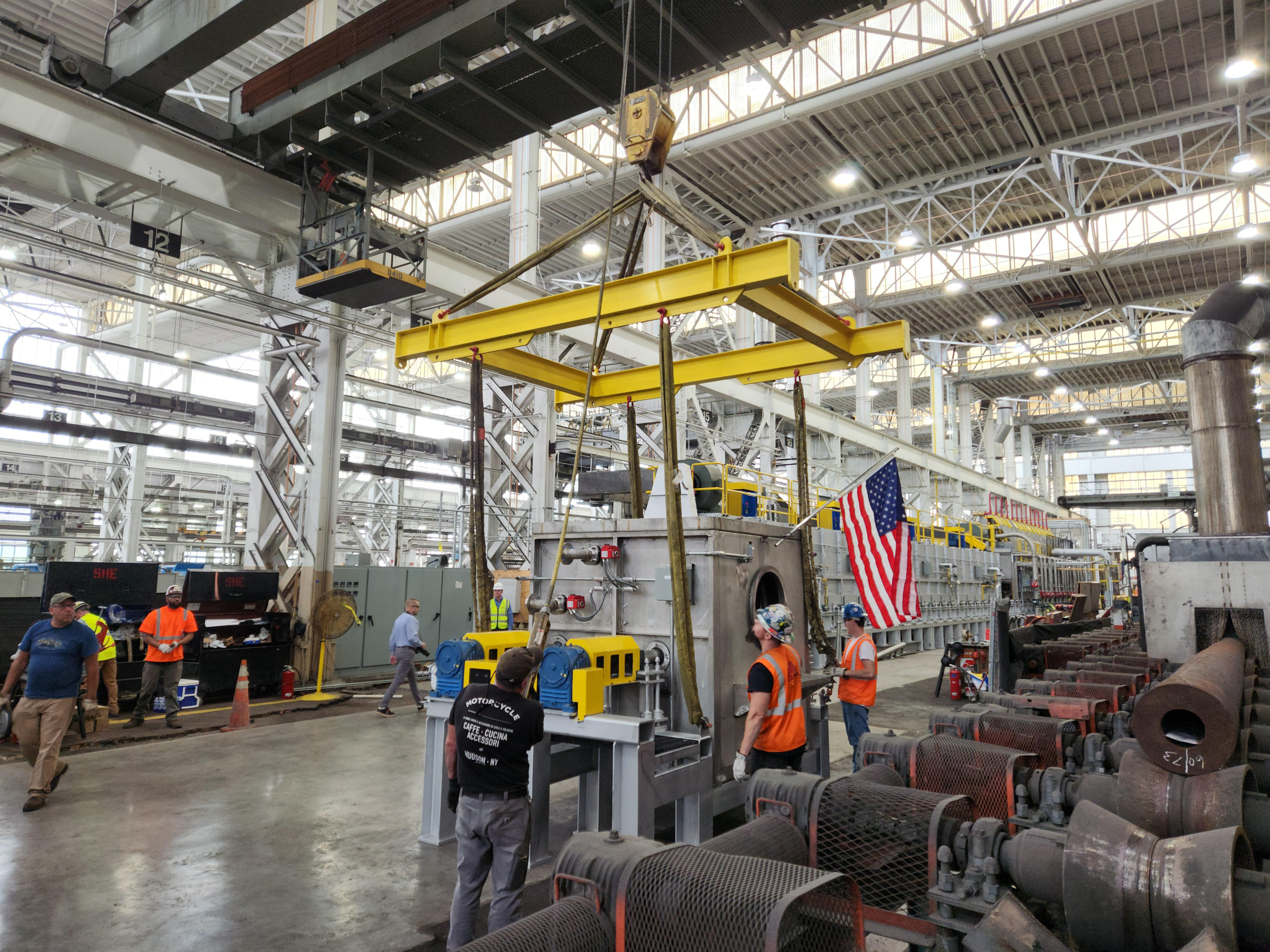 Workers watch a crane lift metal equipment.
