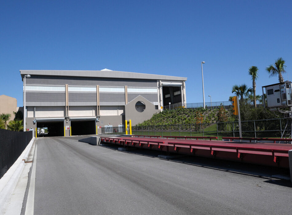 Industrial building, driveway, weighing station, palm trees, clear sky.