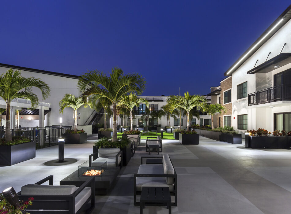 Night courtyard with fire pit, palms, seating, apartments nearby.
