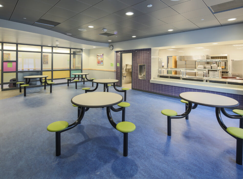 Empty cafeteria with round tables, green stools, blue floor.