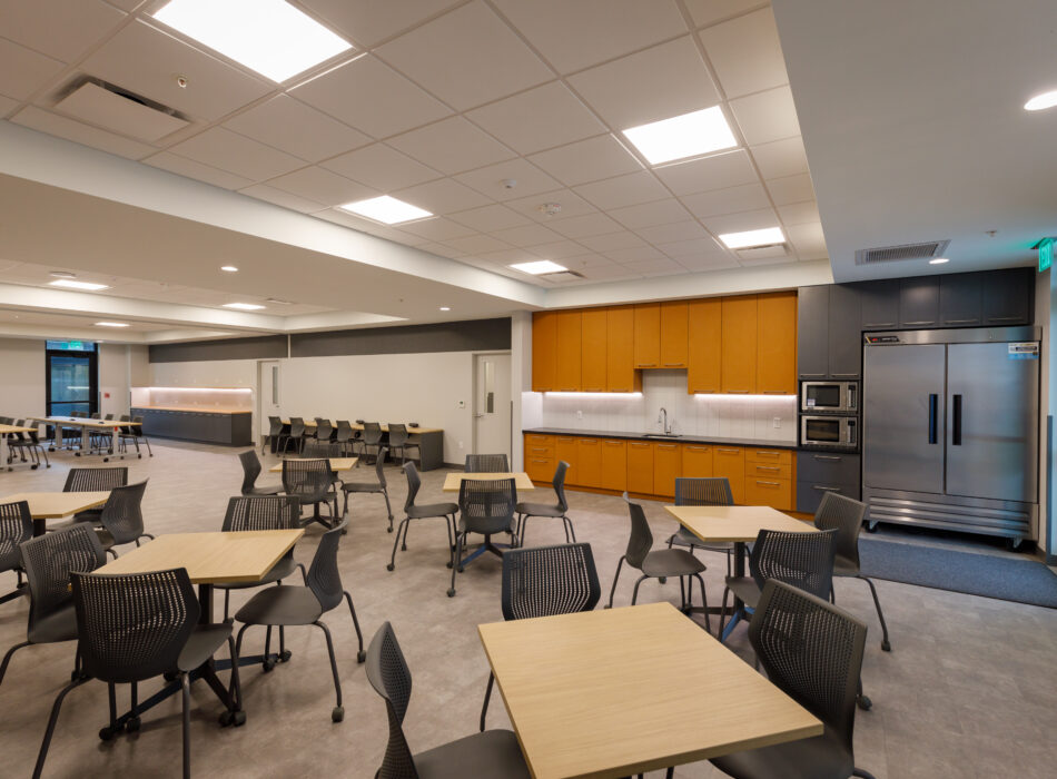 Office break room with tables, kitchenette, and bright lighting.