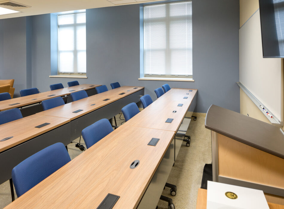 Empty classroom with blue chairs, tables, podium, and screen.