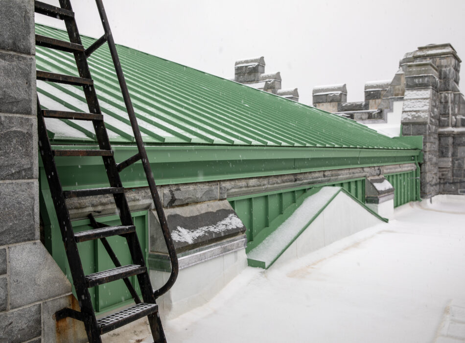 Ladder to snowy green metal roof, stone walls behind.