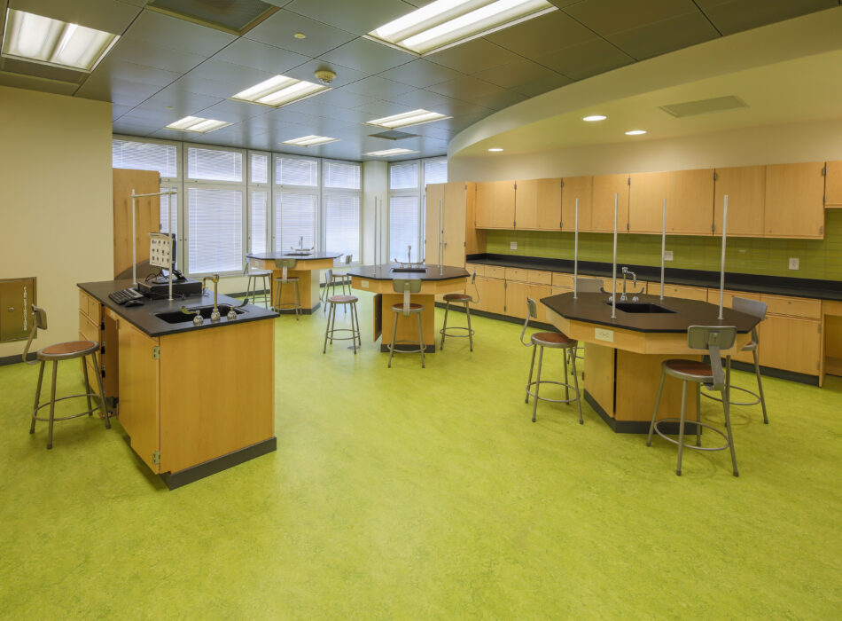 Science classroom with lab tables, stools, computers, green floor.