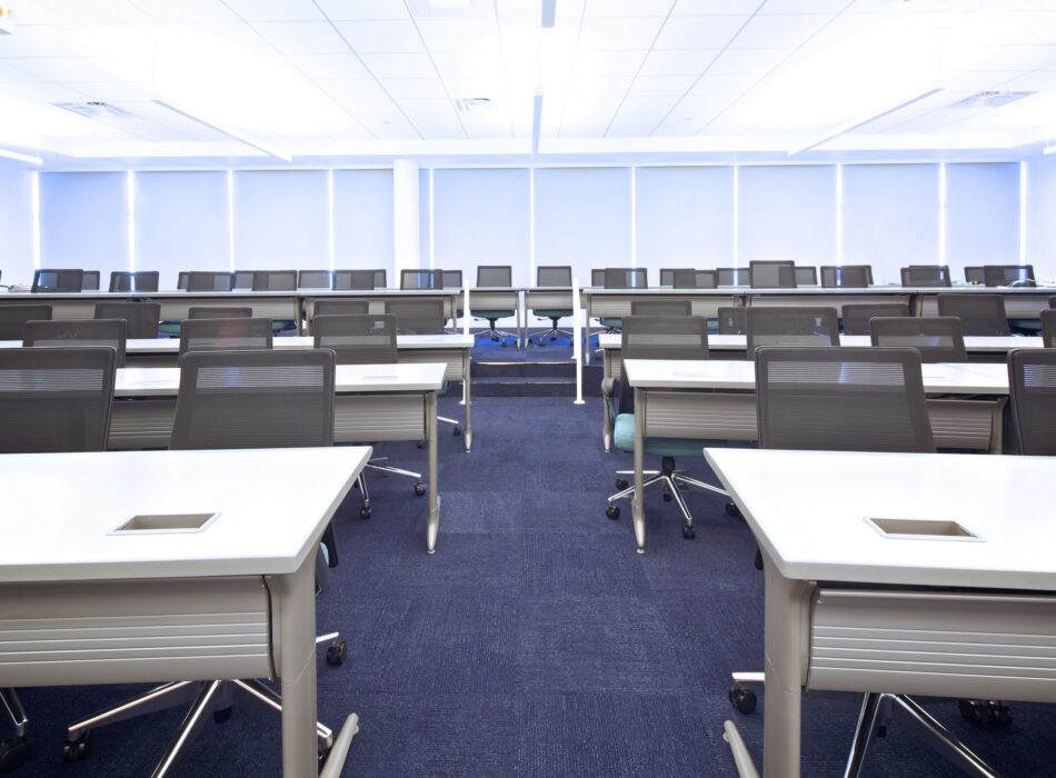 Empty classroom, white desks, black chairs, blue carpet, windows.