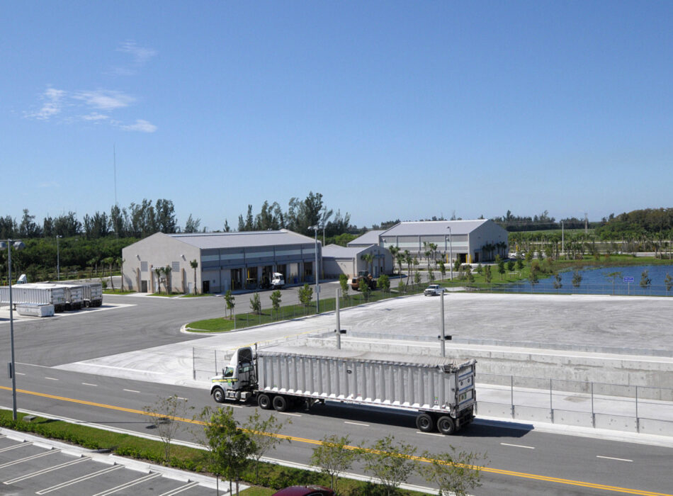Semi-truck passes empty lot, warehouses, and landscaping.
