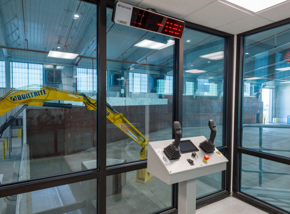 Control panel with joysticks overlooks yellow machine in warehouse.