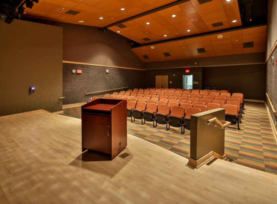 Empty auditorium with orange seats, podium, and warm lighting.