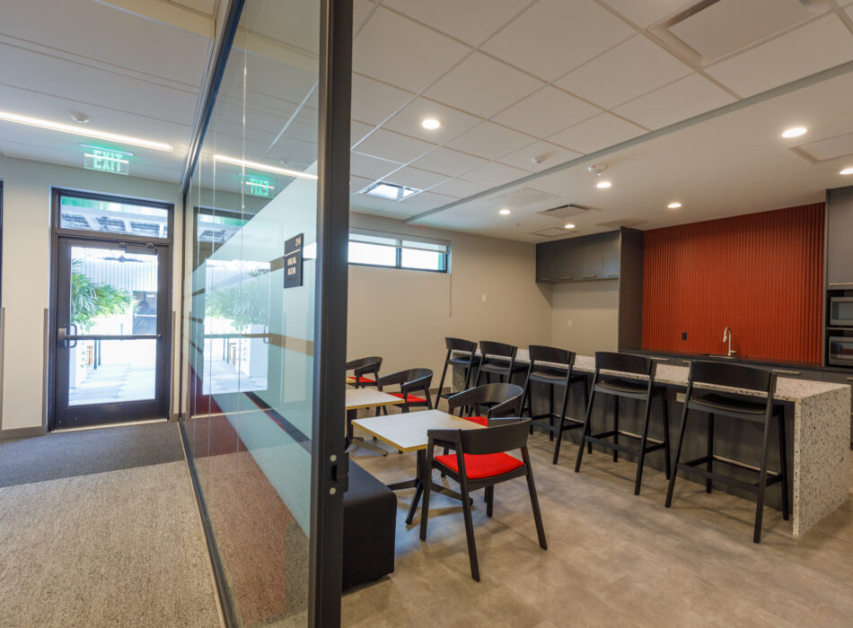 Modern office kitchen and lounge with windows and black chairs.