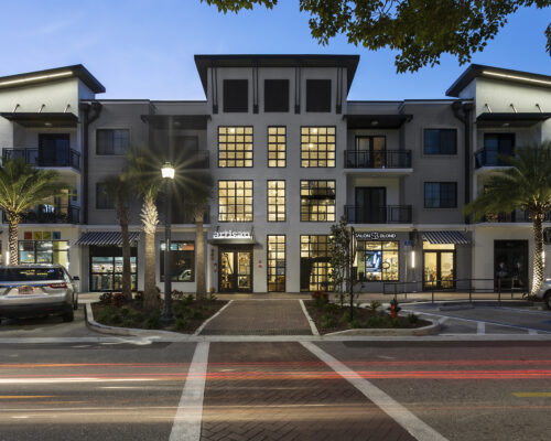 Three-story building with shops, palm trees, dusk traffic.