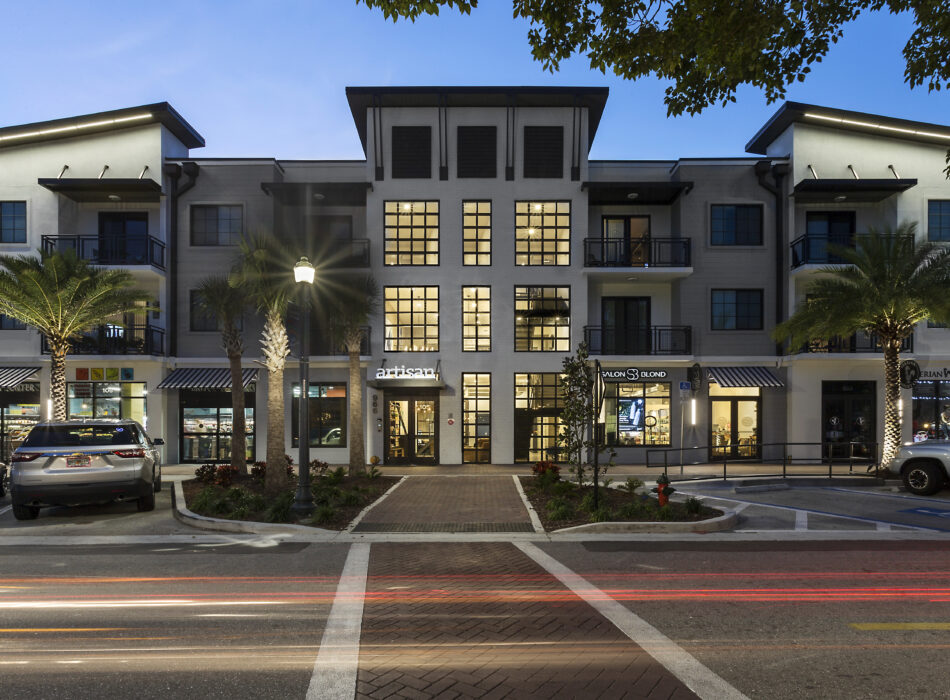 Three-story building with shops, palm trees, dusk traffic.