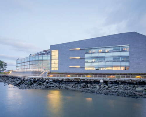Modern building with lit windows beside still water at dusk.