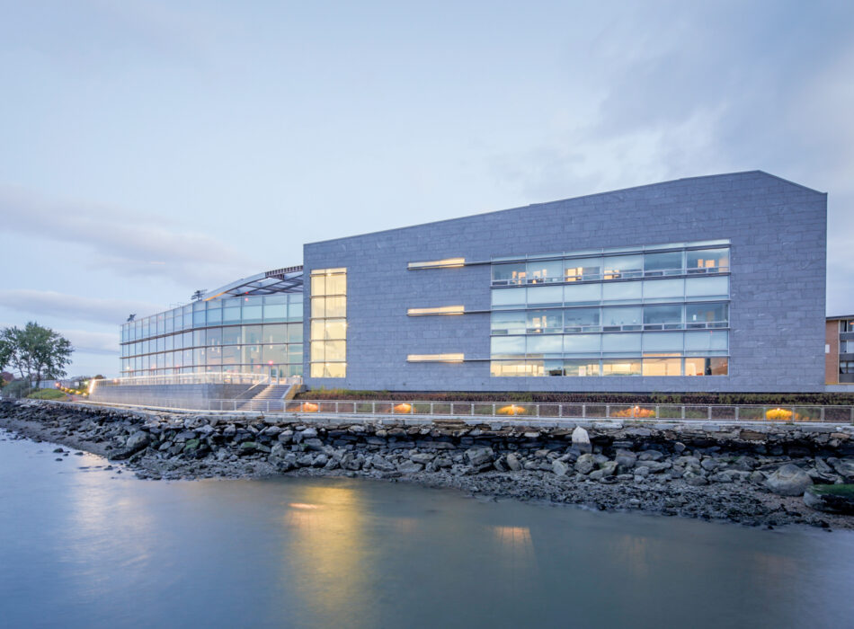 Modern building with lit windows beside still water at dusk.