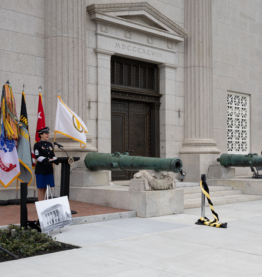Uniformed person at podium outside stone building with flags.