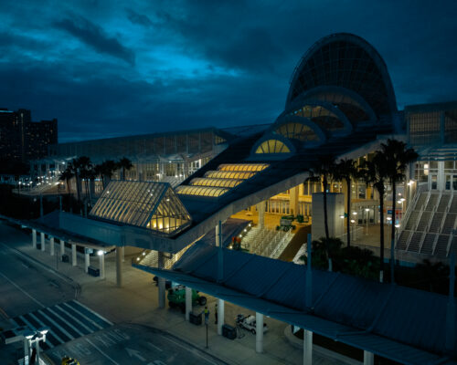Glass convention center at dusk, lights on, palm trees nearby.