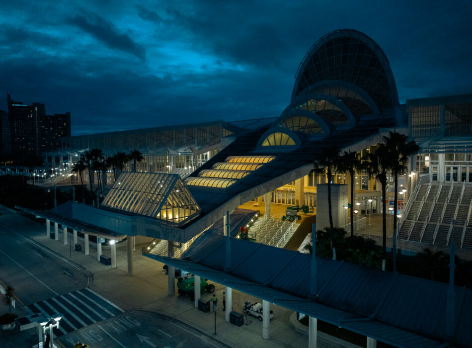 Glass convention center at dusk, lights on, palm trees nearby.