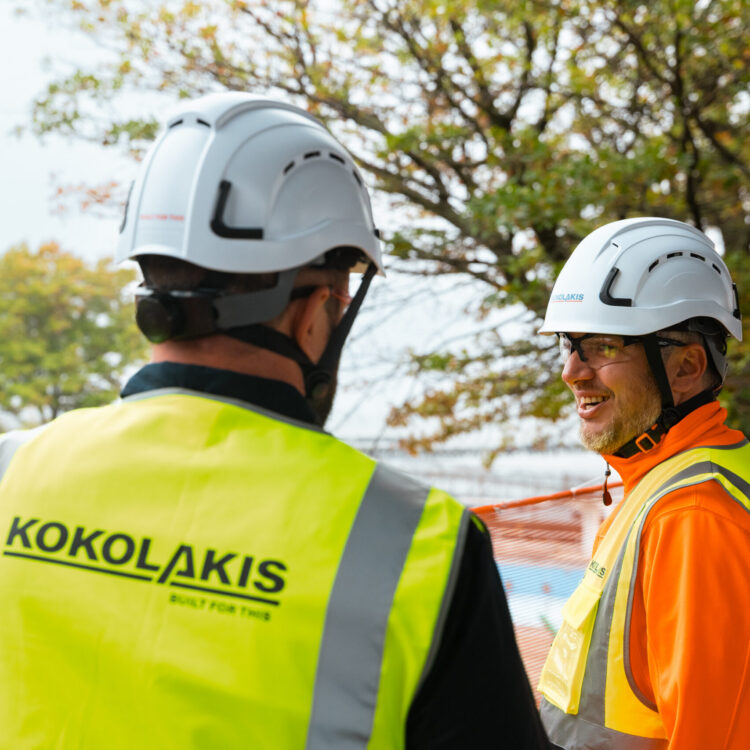 Two construction workers talk at a worksite; vest says Kokolakis.