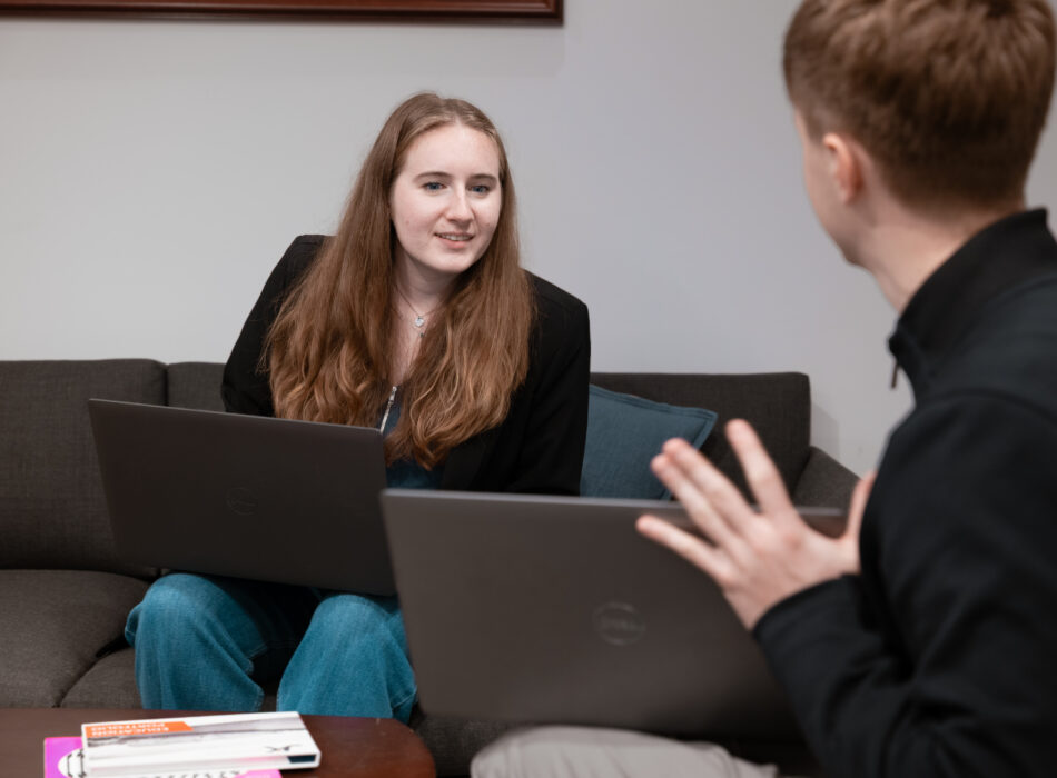 Two people with laptops talk on couch, books on table.
