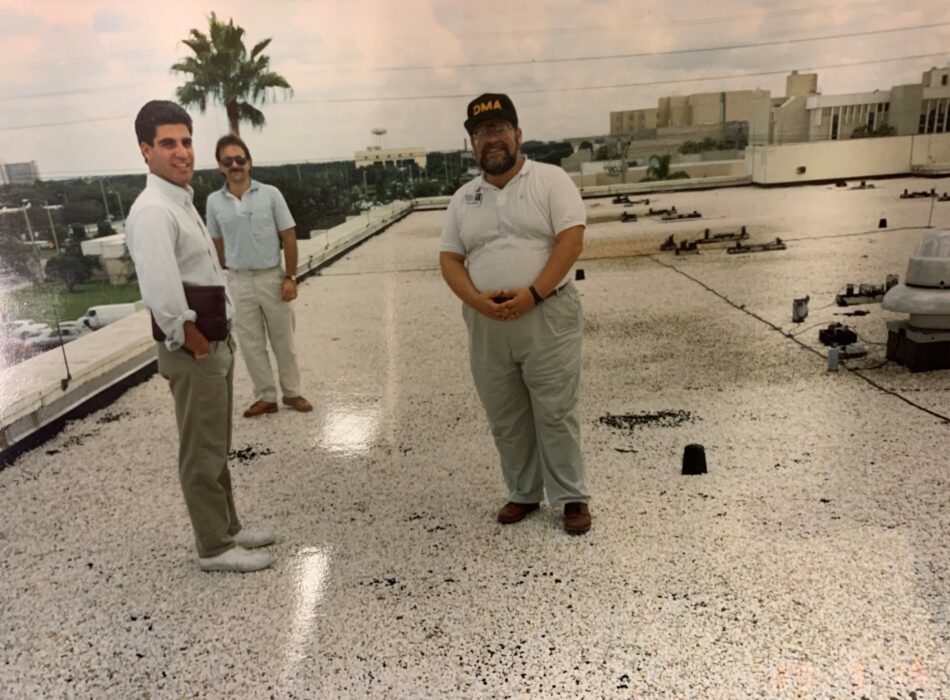 Three men on a gravel rooftop with city background.
