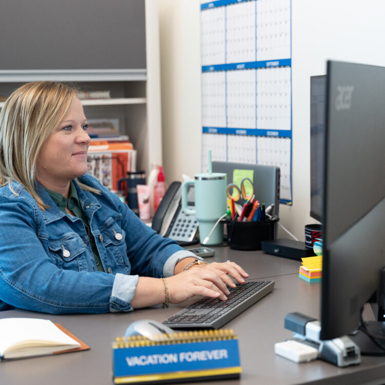 Woman in denim jacket works at desktop, VACATION FOREVER sign.