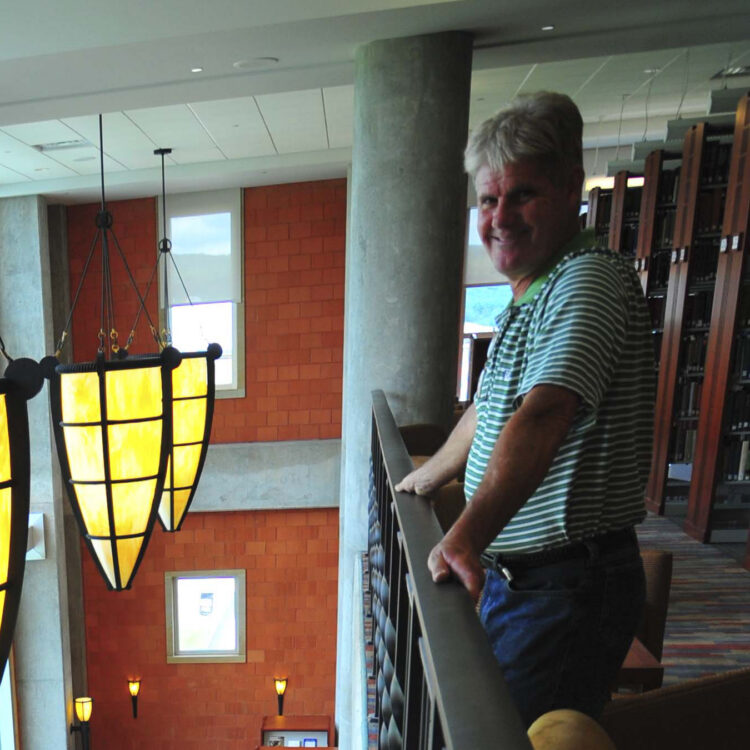 Man in striped shirt on balcony above library with yellow lights.