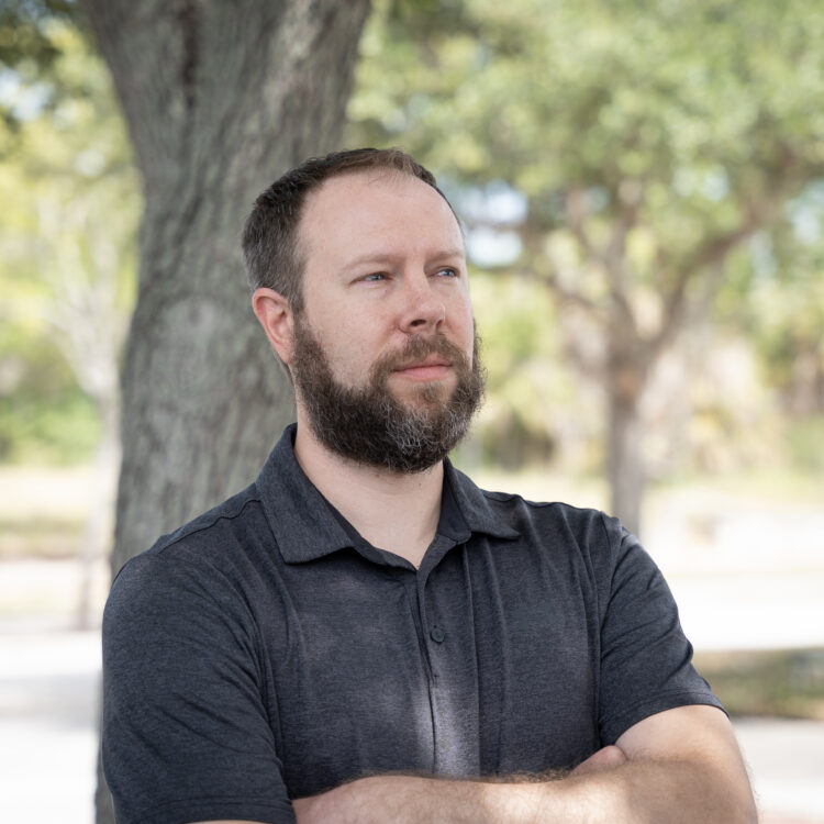 Bearded man in dark polo shirt stands outdoors, arms crossed.